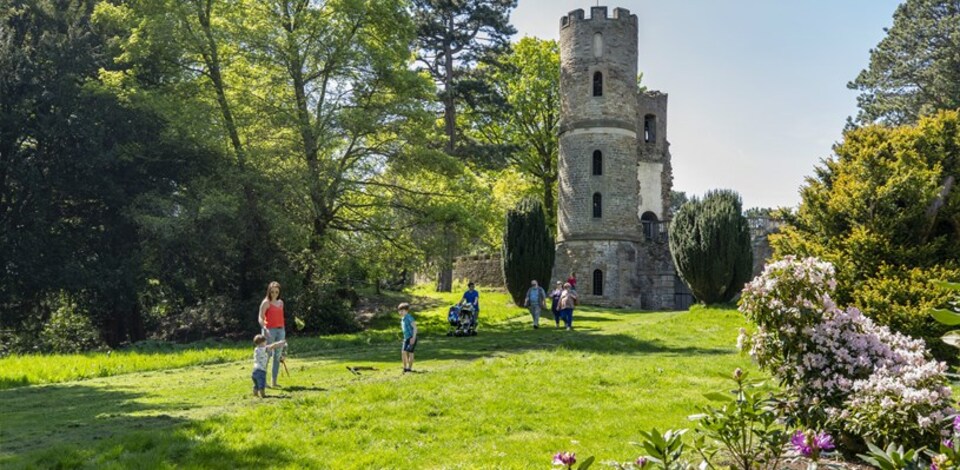 People enjoying a grassy outdoor area near a multi-level stone tower, surrounded by trees, bushes, and flowering plants.